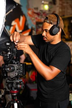 A Black man wearing all black and headphones adjusts the cords on a video camera.