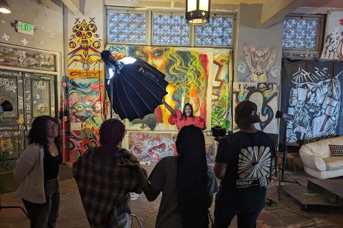 Four members of a film crew face a Black woman in a red dress, who is raising one arm. Behind her, the wall is covered in graffiti.