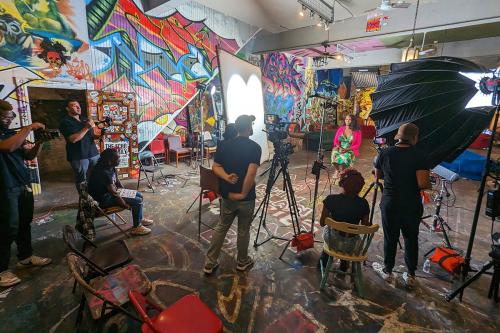 In a large warehouse room covered in colorful graffiti, a film crew with equipment faces a seated interviewee, a Black woman wearing colorful dress and bright pink jacket.