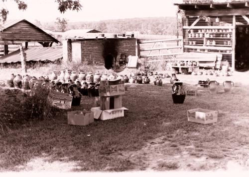 The same scene as the previous photo, but no longer on fire. Instead, finished jugs are lined up in a neat rows in the grass, with several cardboard boxes scattered around too.