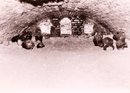 Inside a pottery kiln, with a  low curved brick ceiling and dirt of ash covering the floor. On the sides, a few dark, shining jugs.