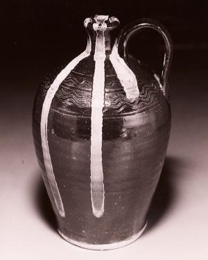 Bulbous dark jug with a single curved handle near the narrow mouth. From the mouth, a design in white glaze seems to ooze out, falling in stripes toward the bottom.