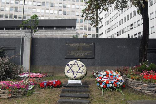 A memorial site, with a flower arrangement of the Star of David in white and blue, plus red flower wreaths on either side, in front of a plaque on a wall. In the background is an office building.