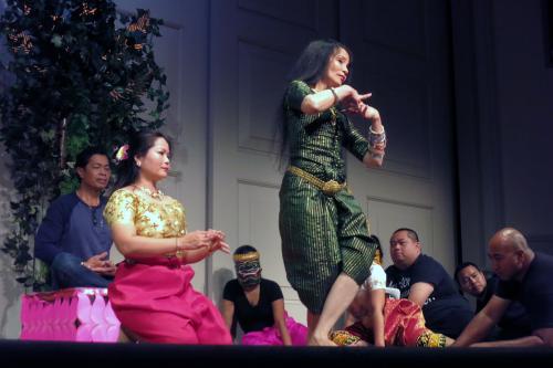 Dancers and musicians rehearse on stage. All are seated except one woman in a dark green dress.