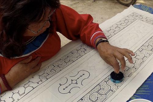 Film still of a woman, seated outdoors, dipping a tiny ink stick in a pot of blue ink as she hand-paints an intricate border design on a white cloth.