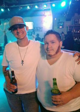 Two people dressing in white T-shirts and each holding a beer bottle, pose arm in arm inside a club, lit by blue stage lights.