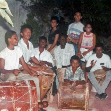 Nine Black people, mostly young men and women and one older man, sit and stand among wooden barrel drums. One blows bubblegum. 