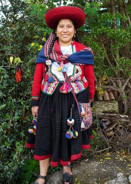 A woman in traditional Peruvian wool dress: black skirt, woven vests and shawl, red wide-brimmed hats all adorned with colorful yarn pom-poms and tassels.