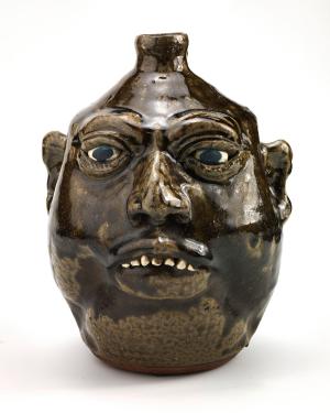 Glazed ceramic jug in the likeness of a human face, with dark skin and white eyes and teeth. The nose, ears, and brow are protruding.