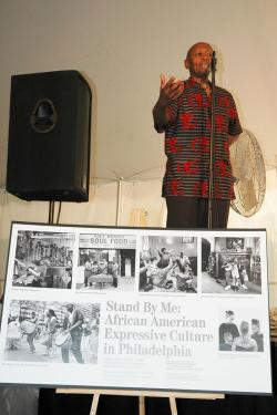 A man speaks into a microphone on stage. In the foreground, a poster display with black-and-white photographs of Black people playing music, gathering, and showing off hairstyles, with the title: Stand By Me: African American Expressive Culture in Philade