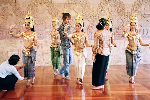 Five women in long skirts and gold headdresses, collars, and belts rehearse a dance in unison.  Three others direct them or adjust their clothing.