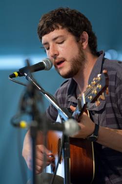 A young man plays guitar and sings into a microphone on stage.