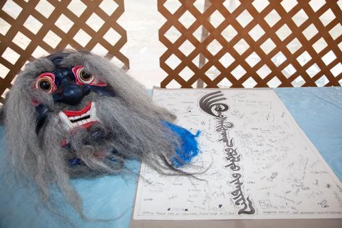 On a display table, a mask with black face, long wispy gray eyebrows and beard, and sharp teeth, next to a page of calligraphy.