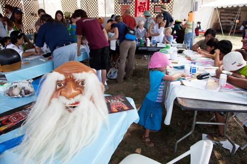 Under an outdoor tent, children and adults draw on long tables. In the foreground, a mask of an old man with long white eyebrows and beard.