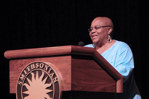 A woman with glasses, gold earrings, and shaved heads speaks at a wooden podium with the Smithsonian sunburst logo on the front.
