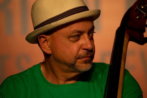A man in a tan fedora and kelly green T-shirt, with a gray goatee and an upright bass beside him, looks to the side out of frame.
