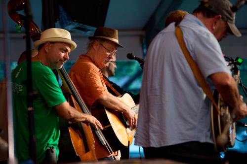 Viewed from the side of an outdoor stage under a tent, a band performs on stage. The instruments visible are upright bass, acoustic guitar, and banjo.