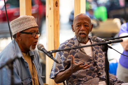 Two men, seated outdoors, speak animatedly into microphones.