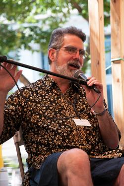 A man with graying hair and beard in a batik print dress shirt and short sits on an outdoor stage, speaking into a microphone.