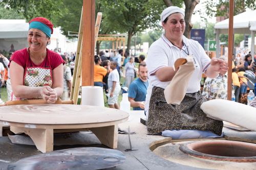 Two women kneel on an outdoor stage forming discs of dough among a crowd. They both laugh.