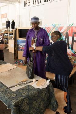 A man and a woman stand behind a table. On the table is a plastic mat and some sharp carving tools. The woman holds a black leather handbag in one outstretched hand and a microphone in the other.