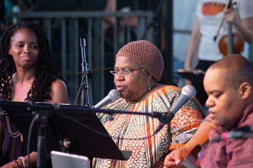 Three women sit on an outdoor stage singing.