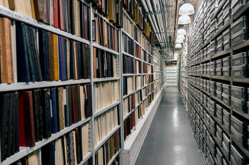 View down the hallway of an archive, with uniform gray boxes on the right and multicolored book or record spines on the left. Pipes are exposed on the ceiling.