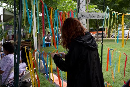 A young woman ties a ribbon onto a piece of twine. Other colorful ribbons blow in the wind, all with writing on them. In the background is a sign that reads: “Chesapeake Collective: Climate Ribbon Project.”