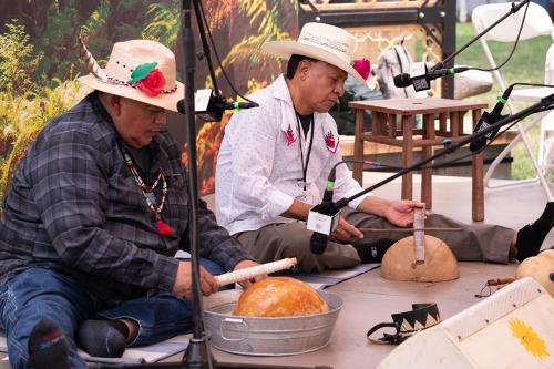 Two men play percussion instruments, seated on an outdoor stage.