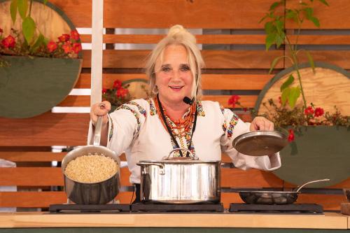 A woman wearing traditional UKrainian embroidered shirt, orange necklaces, and a headset microphone tilts up a pot on a stove to show the brown grains cooking inside.