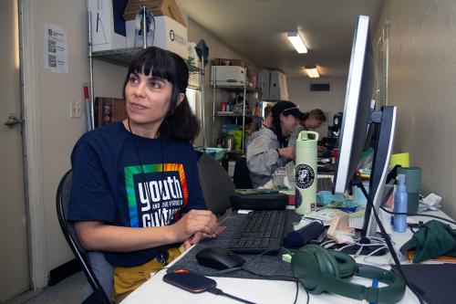 A woman with long dark hair in a ponytail and a navy blue Youth and the Future of Culture T-shirt sits at a computer in a long, skinny room, with others sitting at tables further down.