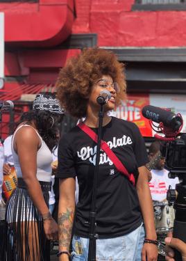 A Black person with an Afro and a Chocolate City T-shirt speaks into a microphone outdoors.
