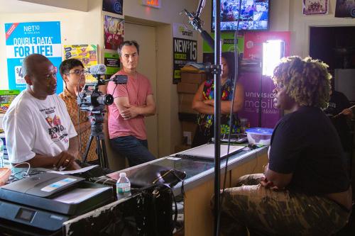 A woman is interviewed on camera, sitting behind a store counter. Three men stand behind the camera, and two other stand to the side.