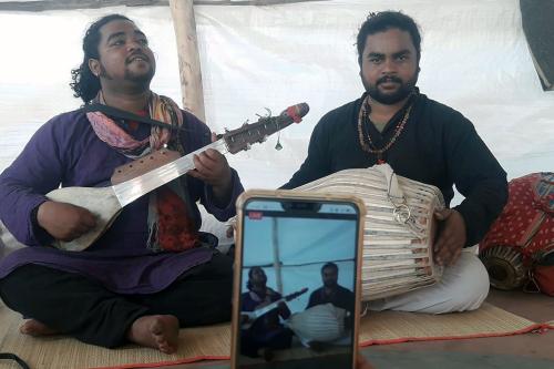 Two brothers play music on a long-necked string instrument and a barrel-shaped drum. An iPhone is in the foreground, filming them.