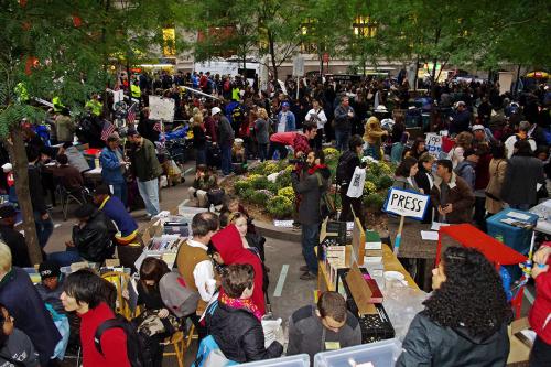 Large crowd of people and protestors in a park. In the foreground is a section of shelves set up and labeled as a library.
