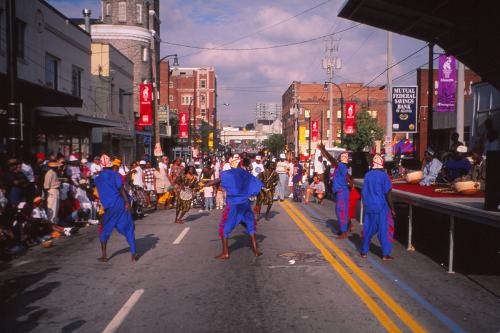 From behind, dancers in matching blue costumes perform in the street for a crowd, alongside a stage.