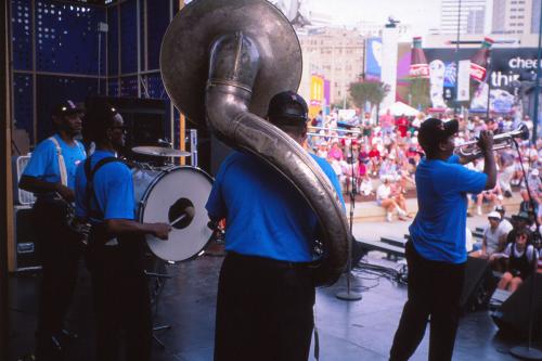Looking out toward the crowd, a brass band in matching blue tees and black pants performs on stage. 