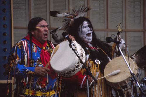 Two Native Americans, one with black and white face paint and feather headdress, the other with long braids, beat on drums and sing on stage. 