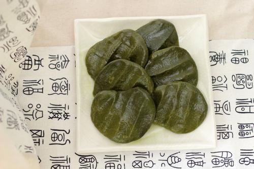 Dark green rice cakes, ridged where finger pressed down to make their shape, on a white share plate, arranged on a printed white cloth.