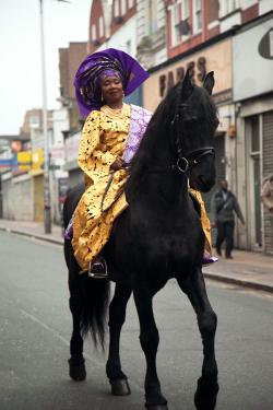 A Black woman couture yellow dress and wide purple headwrap with gold embellishments rides a black horse down a city street.