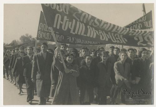 A crowd of people march toward the camera, holding up a banner in the Armenian language. Black-and-white photo, with a watermark for Project Save in the lower right corner.