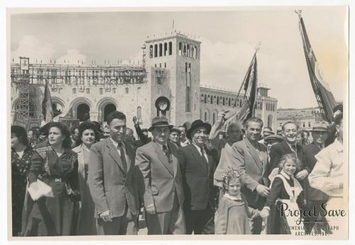 A crowd of men, women, and children, some with flags raised, in formal dress. Behind them, a building is under construction. Black-and-white photo, with a watermark for Project Save in the lower right corner.