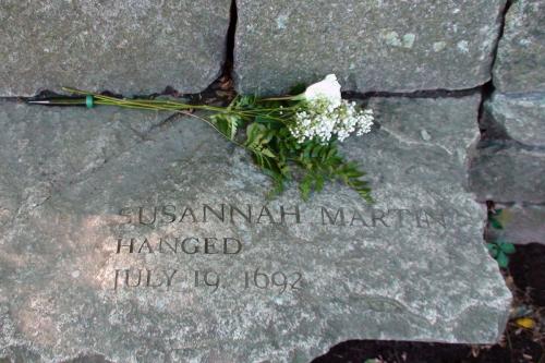 Gray flagstone jutting out from a stone wall, with words etched in: Susannah Martin, hanged July 19, 1692. A bouquet of flowers above the lettering.