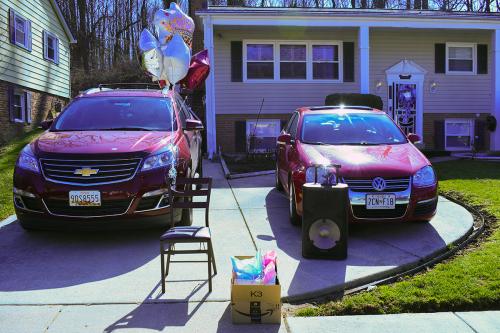 Two parked cars in a driveway in front of a house. In front of them is a chair, with balloons tied to it, and a box of wrapped gifts. 