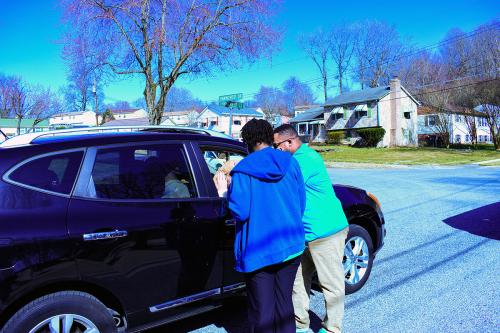 Two people stand in the street, talking with people in a parked car through the passenger-side window.