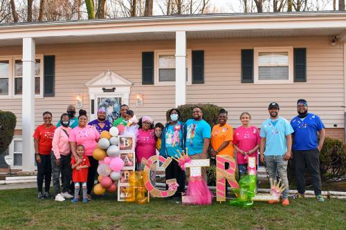 A group of people, some wearing face masks, pose together in front of a home. There are pink and silver balloons and the words BABY GIRL spelled out in paper decorations.