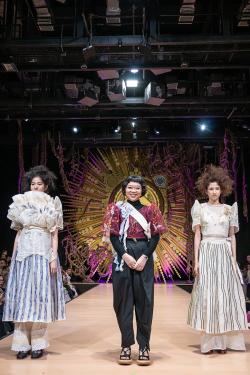 Three people pose on a runway stage. The two on the sides wear white dresses with blue striped skirts, exaggerated sleeves, petticoats, and tall teased-out hair. The woman in the center, the designer, wears black turtleneck and pants with a translucent ma