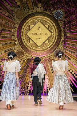 The same three women on the runway as the previous image, now turned and walking away, under an ornate, gold sign that reads TERNOCON.