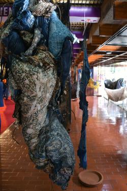 Dyed, printed textile in blue, white, and gray hanging rakishly from the ceiling of an art gallery.