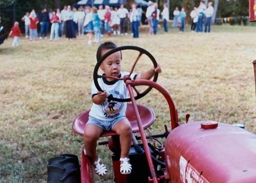 Old color photo of a young boy in the seat of a red tractor, a crowd of people on the field behind him.
